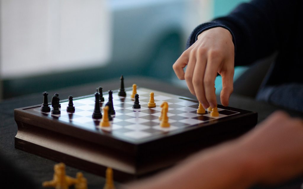 Two people play chess on a wooden board. One players hand hovers over the pieces, contemplating the next move. The board features black and yellow pieces, with a dark background suggesting an indoor setting.