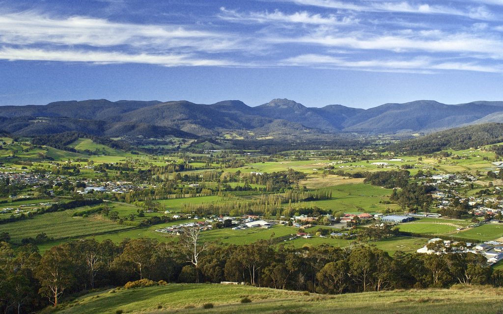 A panoramic view of a lush green valley with scattered houses and farms, surrounded by rolling hills and distant mountains under a blue sky with wispy clouds.