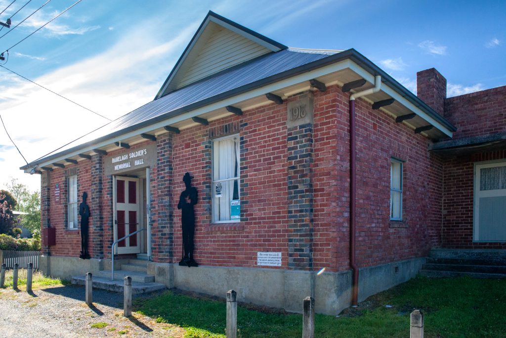 A historic brick building with a metal roof, labeled Nealie Saunders Memorial Hall. Black silhouettes flank the entrance, and a sign with text is visible near the door. The sky is blue with some clouds.