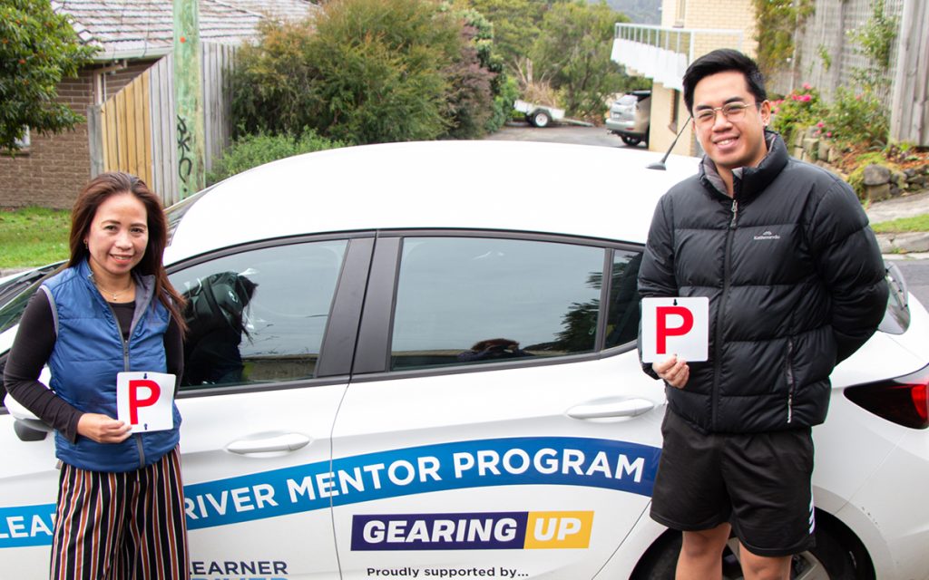 Two people holding red P plates stand beside a car displaying Learner Driver Mentor Program and Gearing Up logos. The scene appears to be in a residential area with trees and houses in the background.