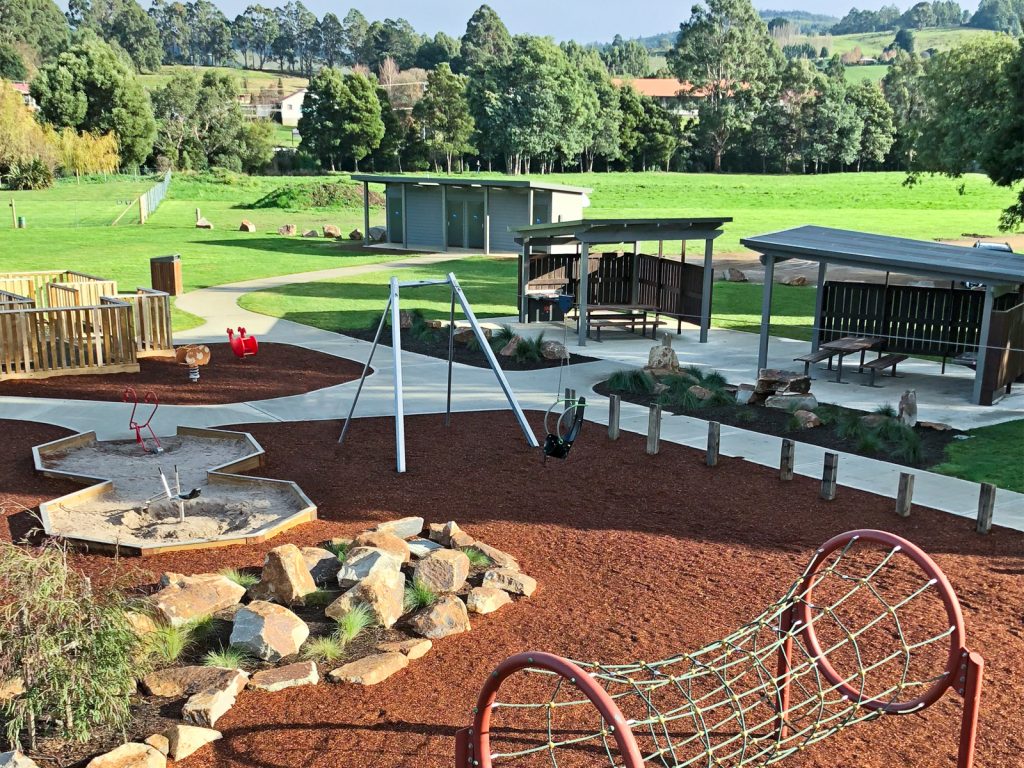 A playground with swings, a climbing net, and seating shelters. Theres a sandbox nearby, set in a landscaped area with rocks and wood chips. Trees and a grassy field are in the background under a clear sky.