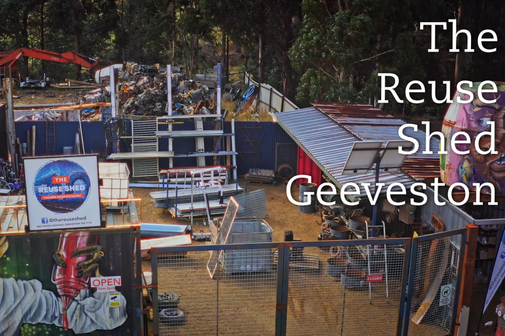 A fenced area filled with stacked metal shelves and assorted items, surrounded by trees. A sign reads The Reuse Shed Geeveston. A large painted mural features a colorful face, and a smaller sign displays the words OPEN and social media handles.