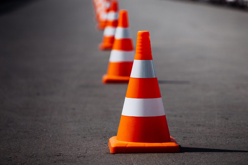 A row of orange and white traffic cones placed along a road, with the cones extending into the distance. The surface of the road appears smooth and is slightly out of focus towards the background.