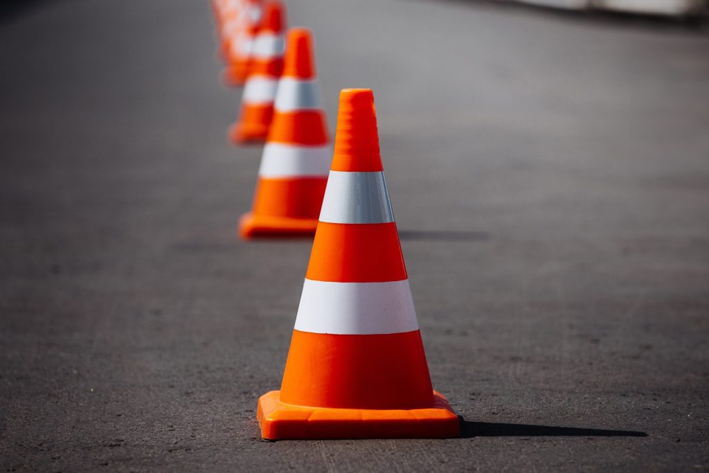 A line of orange and white traffic cones placed on a paved road, extending into the distance. The cones are evenly spaced, with a clear focus on the foremost cone in the foreground. The background is slightly blurred.