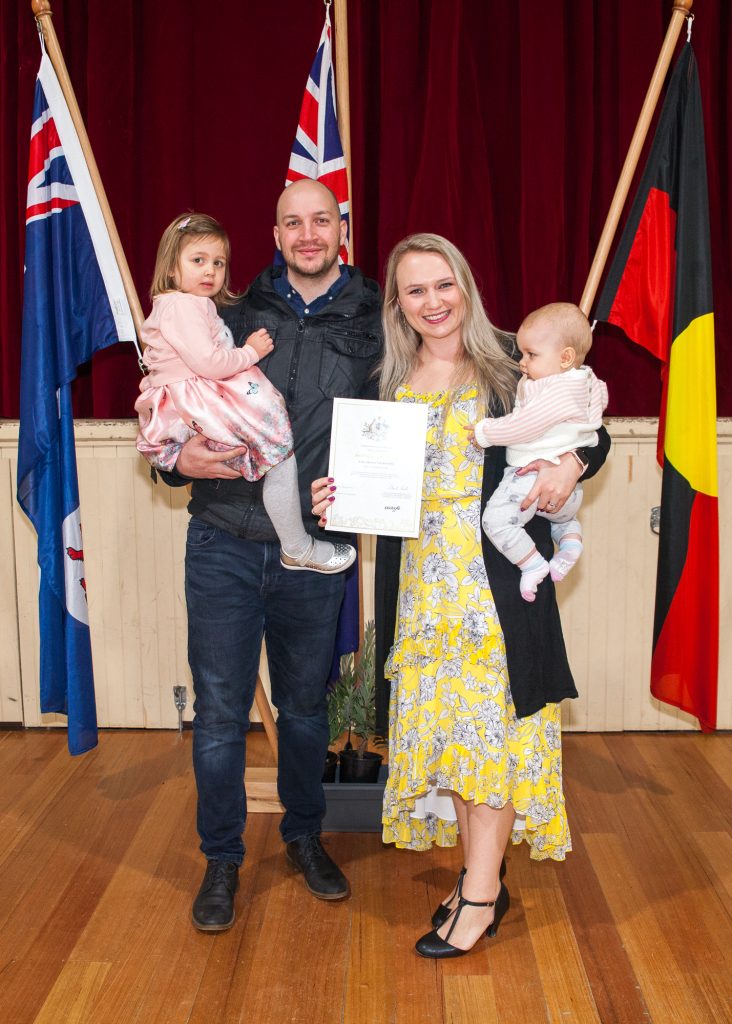 A smiling couple stands indoors holding their two young children. The man holds a daughter in a pink dress, and the woman holds a baby. She is wearing a yellow dress and holding a certificate. Flags are displayed in the background.