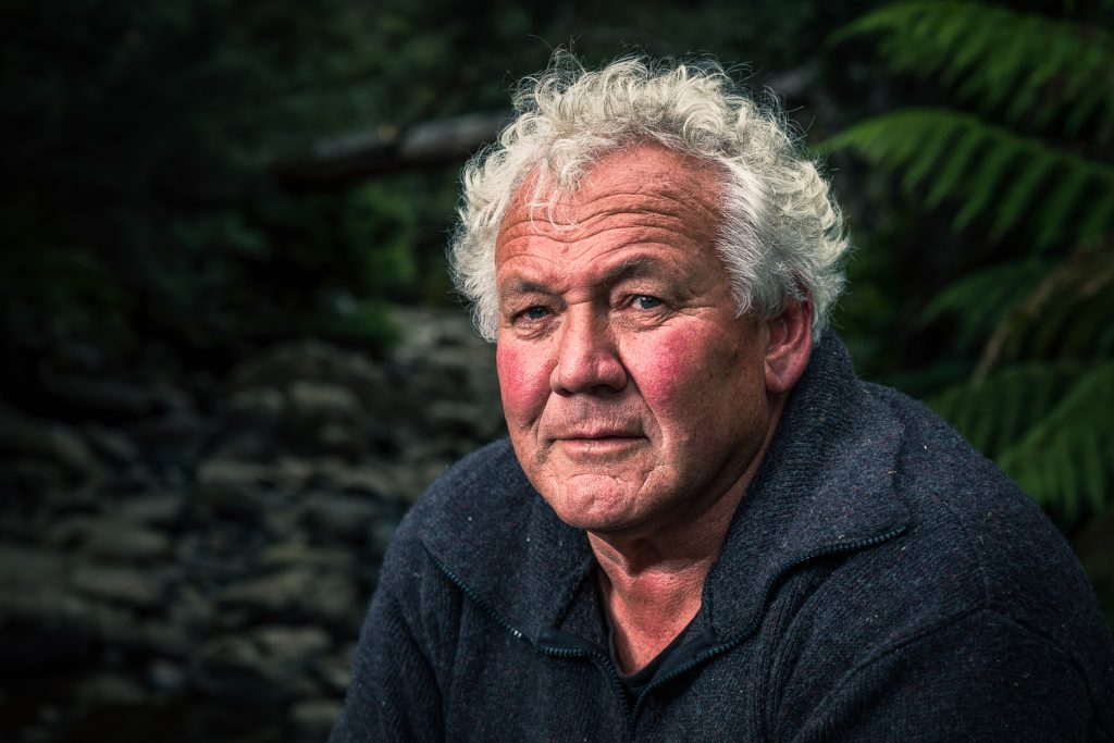 Elderly man with white curly hair and a weathered face, wearing a dark fleece, sits outdoors in a forest setting with ferns and rocks in the background. His expression is thoughtful and serene.