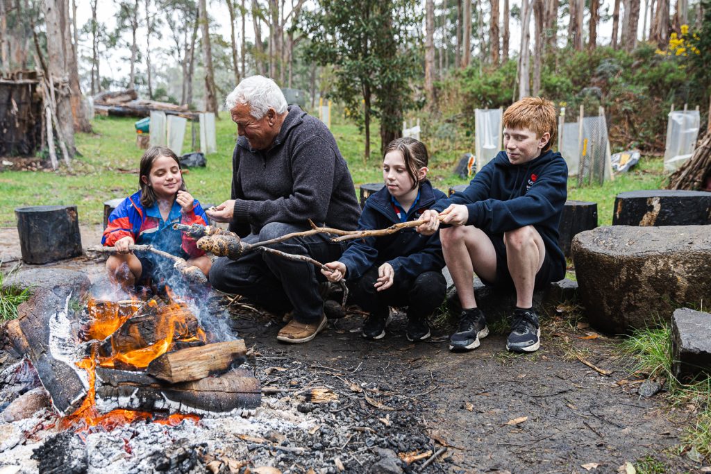 A group of children and an older adult sit by a campfire in a forested area, roasting food on sticks. The background is filled with trees and some protective plant covers. Everyone appears engaged and focused on the fire.