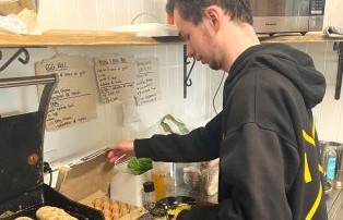 A person wearing a dark hoodie is cooking food on a grill in a kitchen. They are surrounded by shelves with various kitchen items, and there are handwritten notes on the wall.