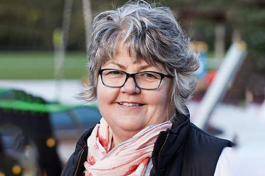 A smiling woman with short gray hair and glasses is wearing a pink scarf and black vest. She stands outdoors with a blurred playground in the background.