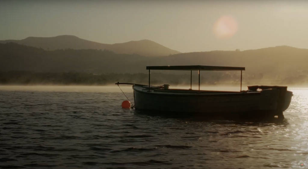 A lone wooden boat floats on a calm lake at sunrise, with mist above the water. The silhouette of distant hills is visible in the background, and sunlight creates a warm, serene atmosphere.