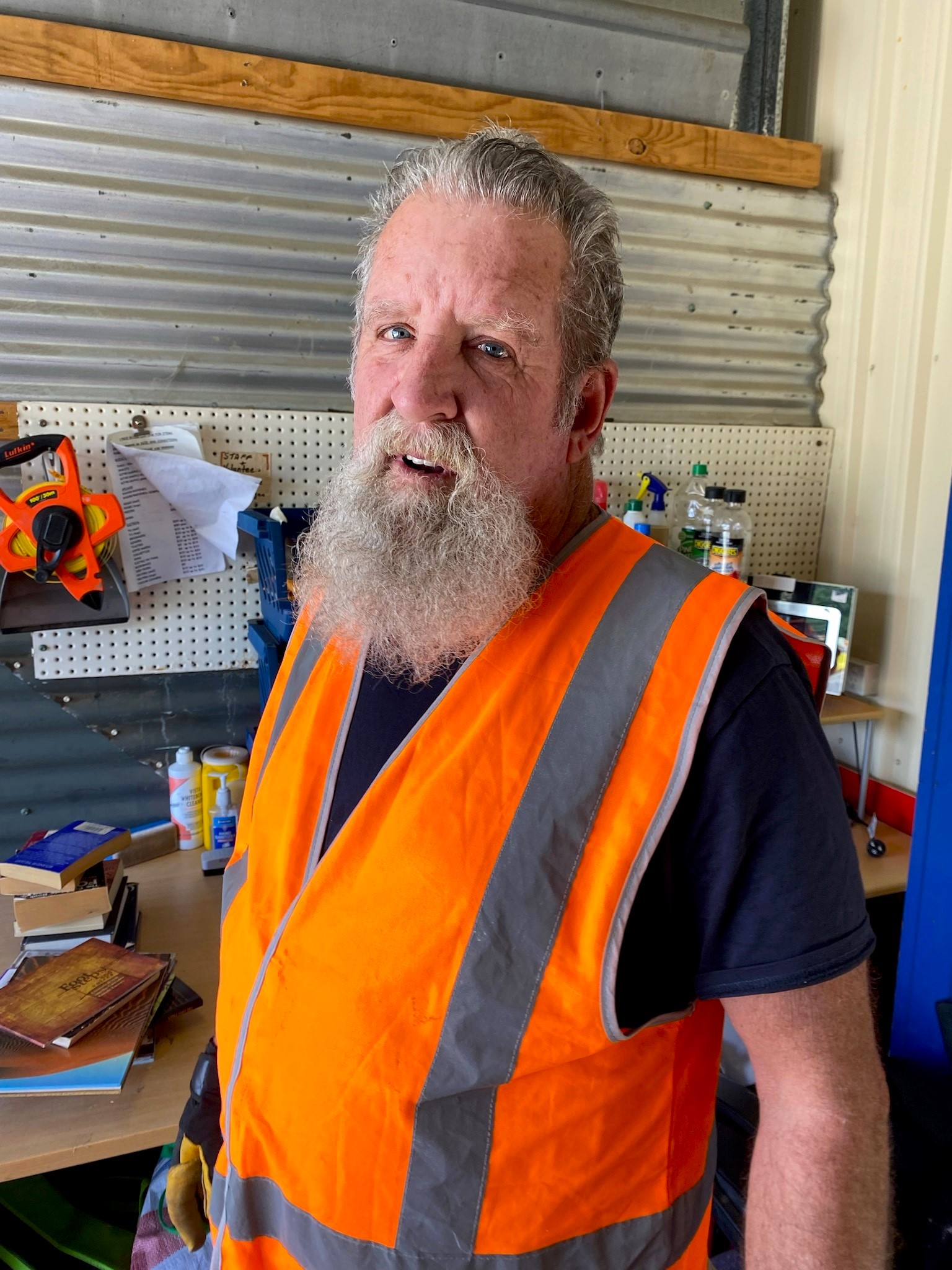 A person with a long white beard wearing an orange safety vest stands in a workshop. Behind them are various tools, books, and supplies on shelves and a pegboard. They are looking toward the camera.