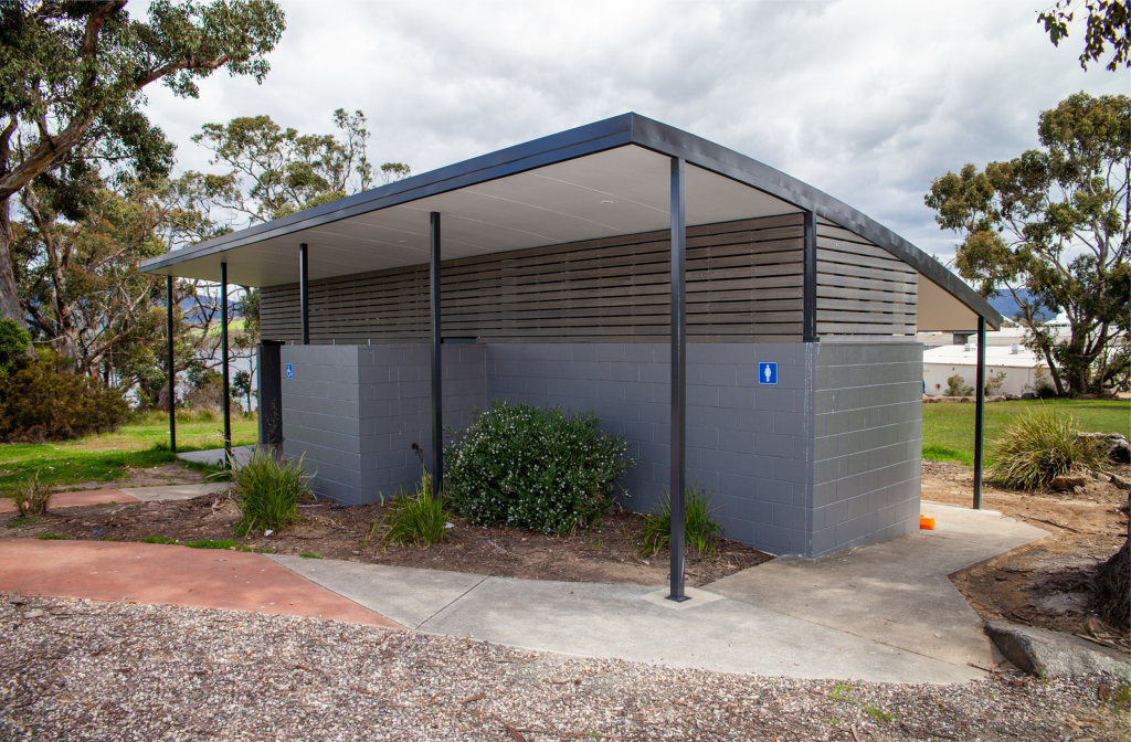 A public restroom facility with a modern design featuring grey brick walls and a metal roof. It is situated outdoors, surrounded by trees and shrubs. There are signs indicating male and female sections on the exterior walls.