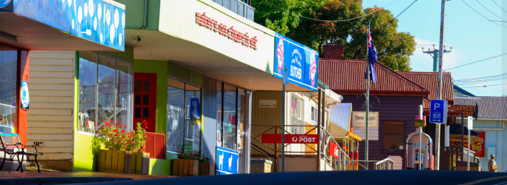 A small town street with colorful shopfronts, including a supermarket and a post office. An Australian flag is visible. The pavement is deserted, and sunlight casts shadows from nearby trees and buildings.
