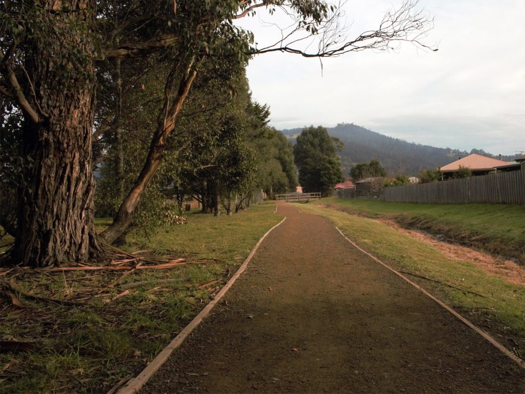A dirt path lined with trees and grass leads into the distance, flanked by a wooden fence on one side. Hills covered in trees are visible in the background under a cloudy sky.