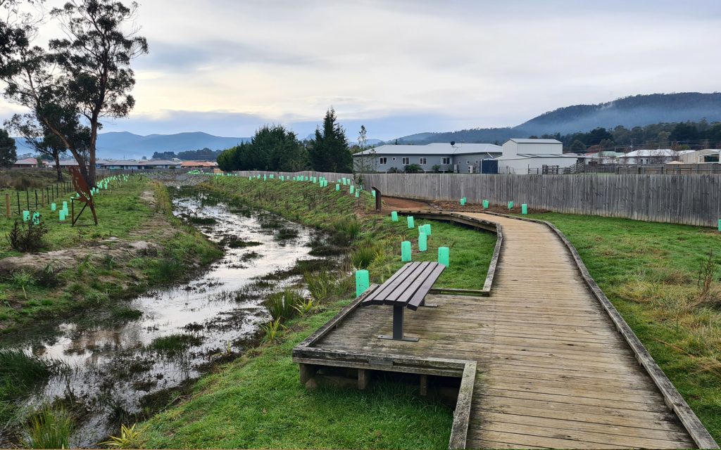A wooden boardwalk runs alongside a narrow stream in a grassy area with young trees in protective tubes. Residential buildings and mountains are visible in the background under a cloudy sky. A lone bench is placed on the boardwalk.