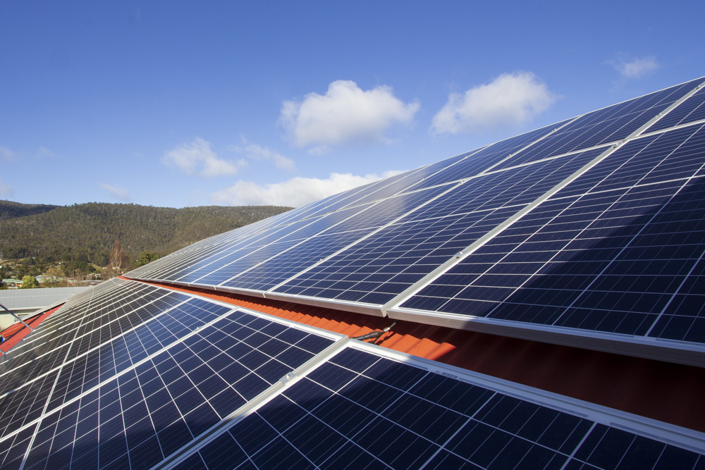 Solar panels on a red roof under a blue sky with scattered clouds. The panels are angled towards the sun, and a tree-covered hillside is visible in the background.
