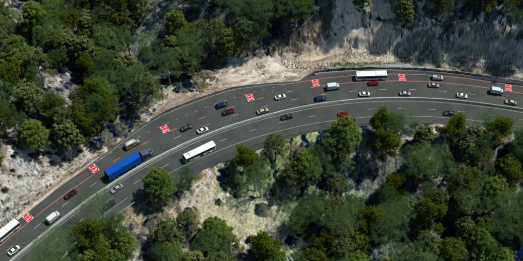 Aerial view of a winding road through a forested area, with various vehicles including cars, trucks, and buses traveling in both directions. Traffic signs and lanes are visible, and the surrounding landscape is dotted with trees and rocky terrain.