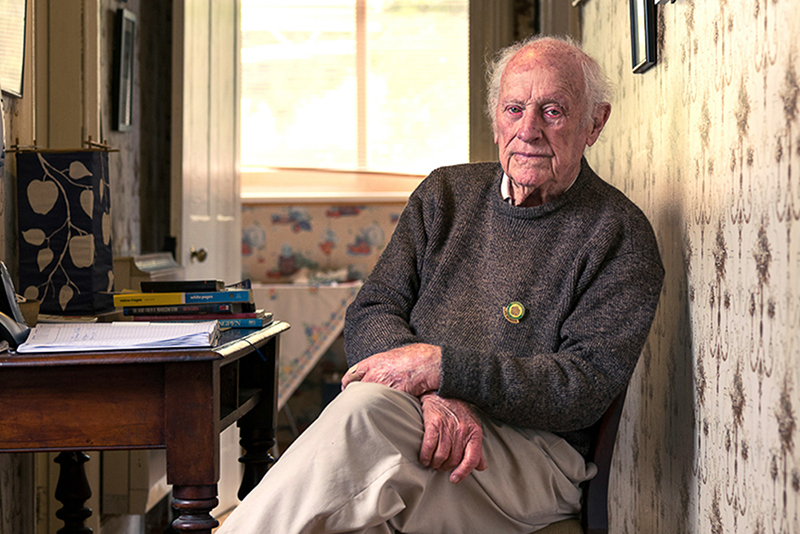An elderly man with white hair sits on a chair, leaning against a patterned wall. He wears a brown sweater and beige pants. A small table beside him holds books and papers. The room is softly lit by a window in the background.