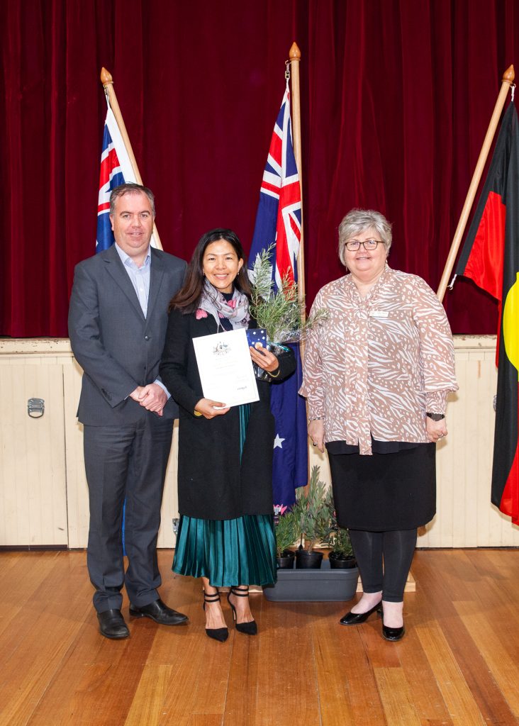 Three people stand on a wooden floor in front of Australian and Indigenous flags. The woman in the middle holds a certificate and smiles. The man and woman flanking her are dressed formally, with the woman on the right wearing glasses.