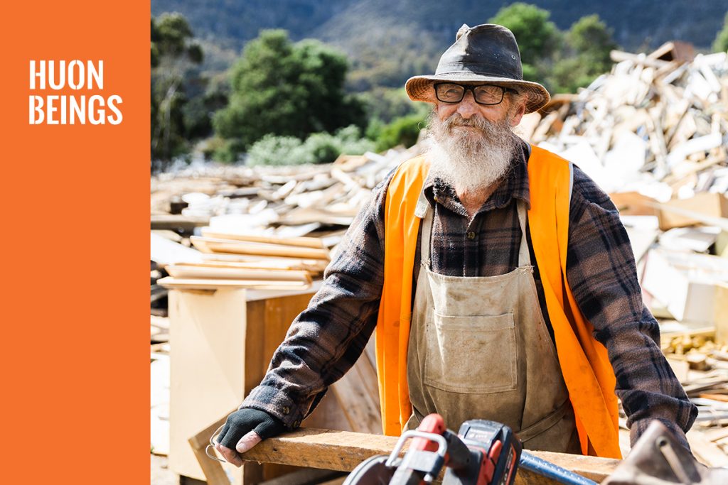 Elderly man with a bushy white beard, wearing a hat, glasses, orange safety vest, and plaid shirt, stands amidst a pile of debris. He smiles slightly while leaning on a piece of wood. The text HUON BEINGS is visible on the left side against an orange background.