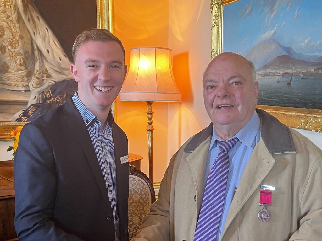 Two men smiling and shaking hands in a warmly lit room with elegant decor. One man wears a suit and name tag, while the other wears a coat and tie with a medal. A lamp and paintings are visible in the background.