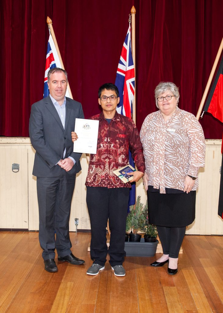 Three individuals stand on a wooden floor in front of a red curtain, with Australian flags behind them. The person in the middle holds a certificate and a small box. The individuals on either side wear formal clothing and smile at the camera.