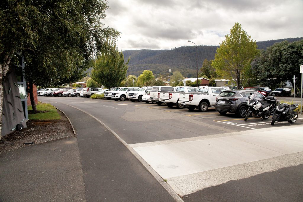 A parking lot filled with various cars and a motorcycle on a cloudy day. Trees line the sidewalk on the left, and hills are visible in the background. The sky is overcast, giving a muted light to the scene.