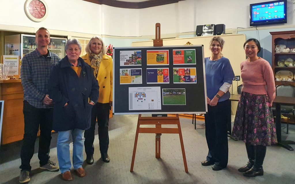 Five people standing around a display board on an easel showing various colorful posters and papers. They are in a room with a bar area and a TV on the wall. Tables and chairs are in the background. Everyone is smiling.