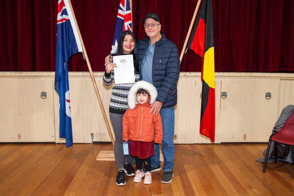 A smiling woman holds a certificate, standing next to a man and a child in a red coat with a fuzzy hood. Theyre in front of Australian and Aboriginal flags against a red curtain backdrop on a wooden floor.