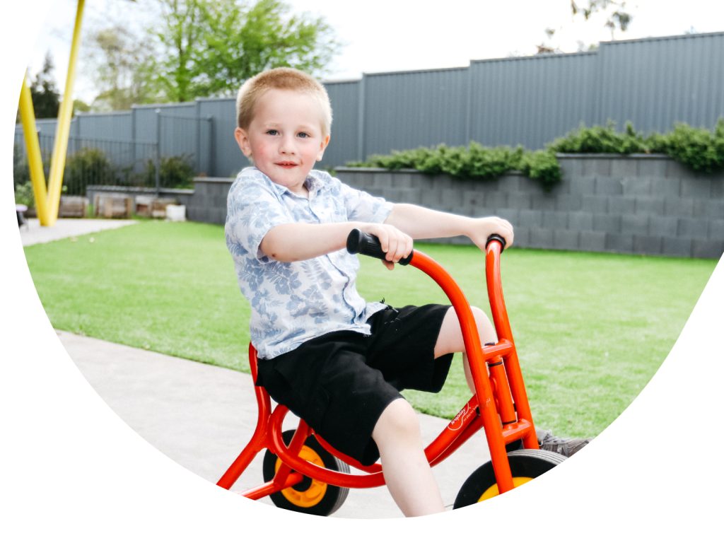 Young child with short hair rides a bright orange tricycle on a paved path next to a green lawn. The child wears a light blue patterned shirt and black shorts. The background features a gray fence and greenery.