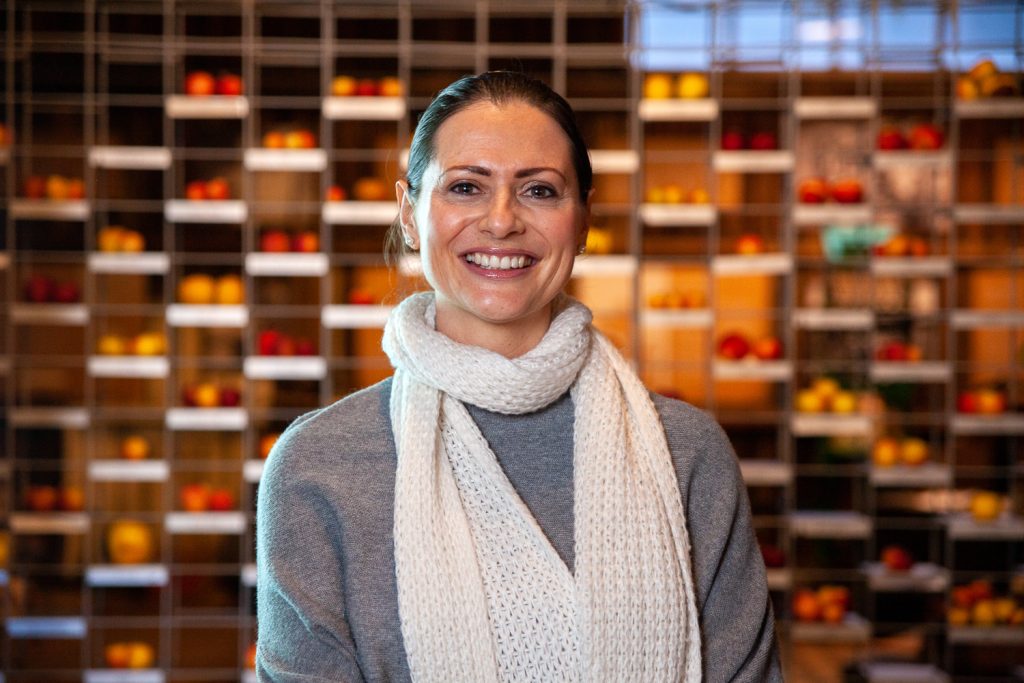 Woman smiling, standing in front of a wall of shelves filled with colorful fruit. She wears a light scarf and a grey sweater. The background is softly blurred, highlighting the vibrant oranges and reds of the fruit.