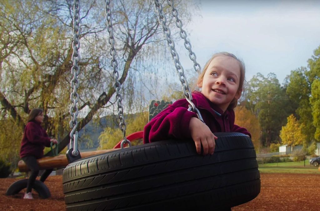 A young child with brown hair and wearing a maroon top happily swings in a tire swing at a playground. Another person in a matching outfit stands nearby. Trees and greenery are visible in the background.