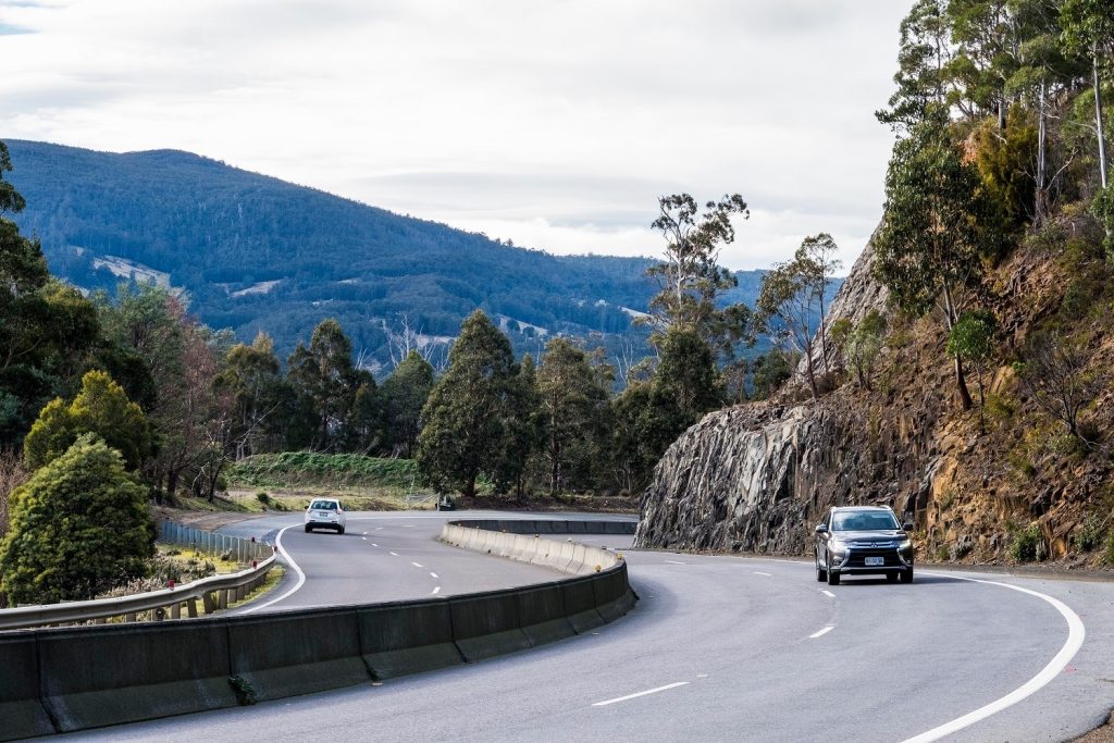 A winding road with two cars driving, surrounded by rocky cliffs and dense green forest. Overcast skies hang above rolling hills in the distance.