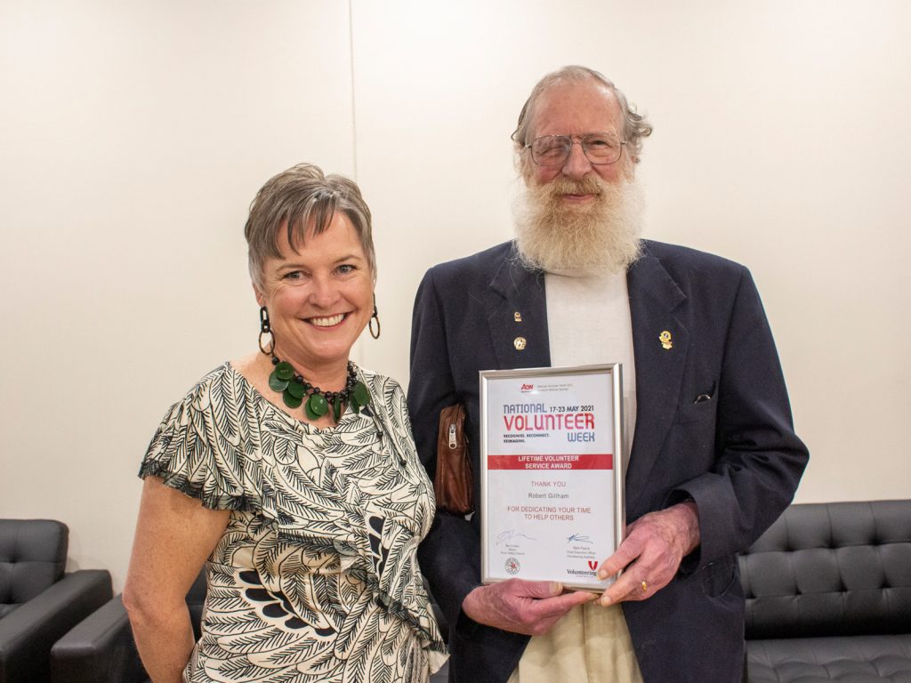 A smiling woman and an older man with a white beard stand together indoors. The man is holding a framed certificate recognizing his volunteer work. They are in a room with black leather sofas in the background.