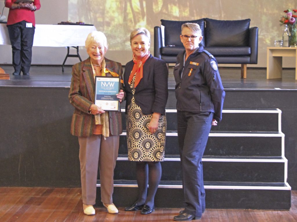 Three women stand on a stage. One holds a certificate. They are smiling and posed in front of a dark sofa and a table. The woman on the left wears a striped jacket, the center in a patterned dress, and the right in a uniform.