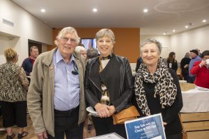 Three people smiling at an indoor event, standing in front of a table. The man on the left wears glasses and a blue shirt. The two women are wearing jackets, with the woman on the right wearing a leopard print scarf. Other people are in the background.