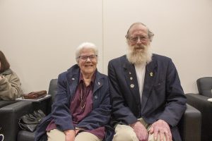 Elderly couple sitting on a black couch, smiling. Both wear dark blazers; the woman is in glasses and a purple top, while the man has a long white beard. A purse and another person are partially visible on the left.