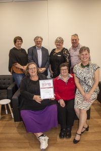 Seven people are gathered in a room with a gray couch and chair. Four are seated, and three are standing. The person seated in the front holds a framed certificate. They are all smiling at the camera.
