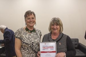 Two smiling women in a room pose with a volunteer service award. One woman holds a framed certificate that reads National Volunteer Week. Other people and a couch are visible in the background.
