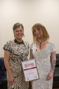 Two women stand together indoors, smiling. One holds a framed certificate for National Volunteer Week. The woman on the left wears a patterned dress, and the woman on the right wears a light dress and has a colorful tattoo on her arm.