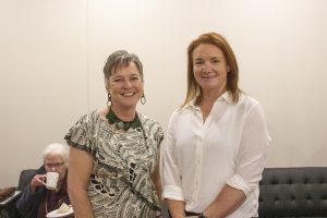 Two women smiling at the camera; one wearing a patterned dress and a necklace, and the other in a white shirt. In the background, an older person is sitting on a couch, holding a cup.