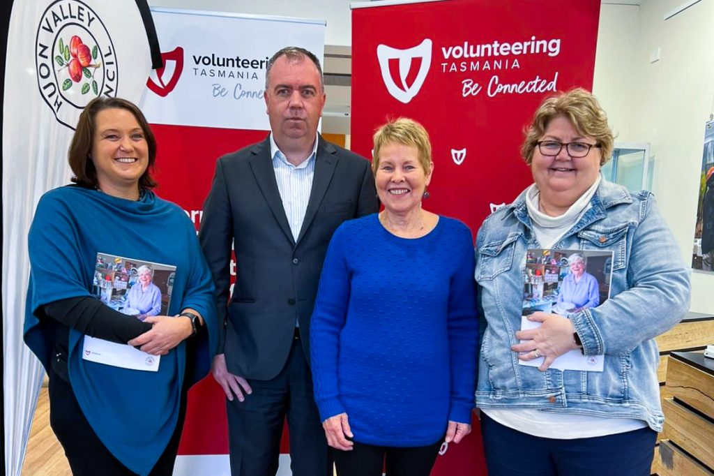 Four people stand smiling in front of banners for Volunteering Tasmania. Three women hold brochures while a man stands with his hands at his sides. They are standing on wooden flooring in a room with light-colored walls.