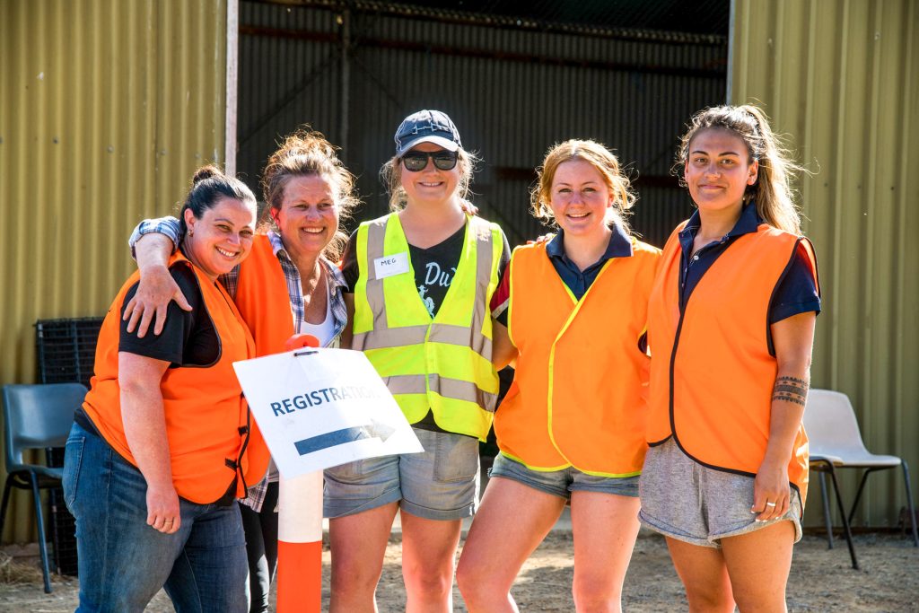 Five smiling people wearing orange safety vests stand together outdoors. One person holds a Registration sign. They are in front of a corrugated metal wall with chairs scattered around. The scene appears sunny and bright.