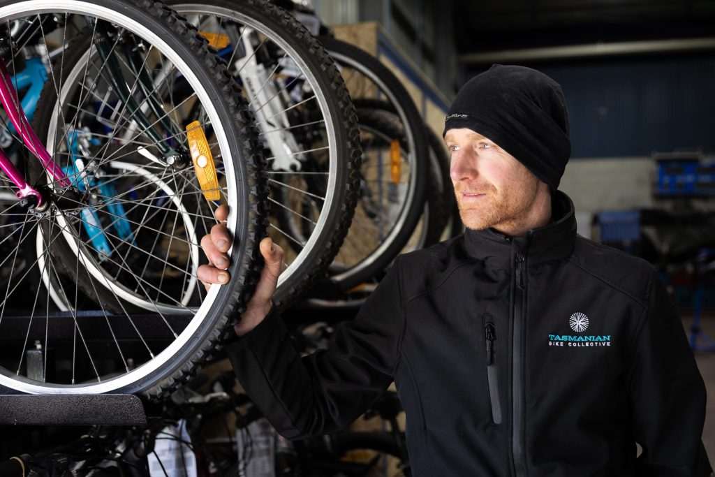 A person in a black jacket and hat inspects a bicycle wheel in a workshop filled with various bikes. They have light facial hair and are looking to the right. The scene is brightly lit, highlighting the bikes and the persons thoughtful expression.
