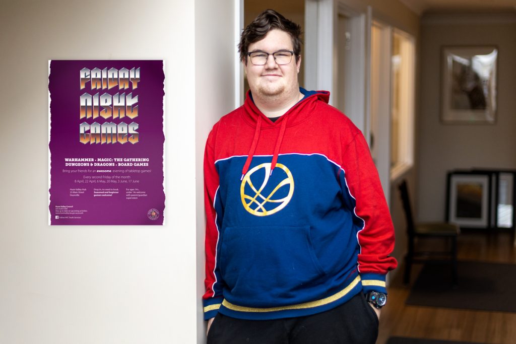 A person wearing glasses and a red and blue hoodie stands indoors near a purple poster that reads Friday Night Games. The poster mentions Warhammer, Magic: The Gathering, Dungeons & Dragons, and board games.