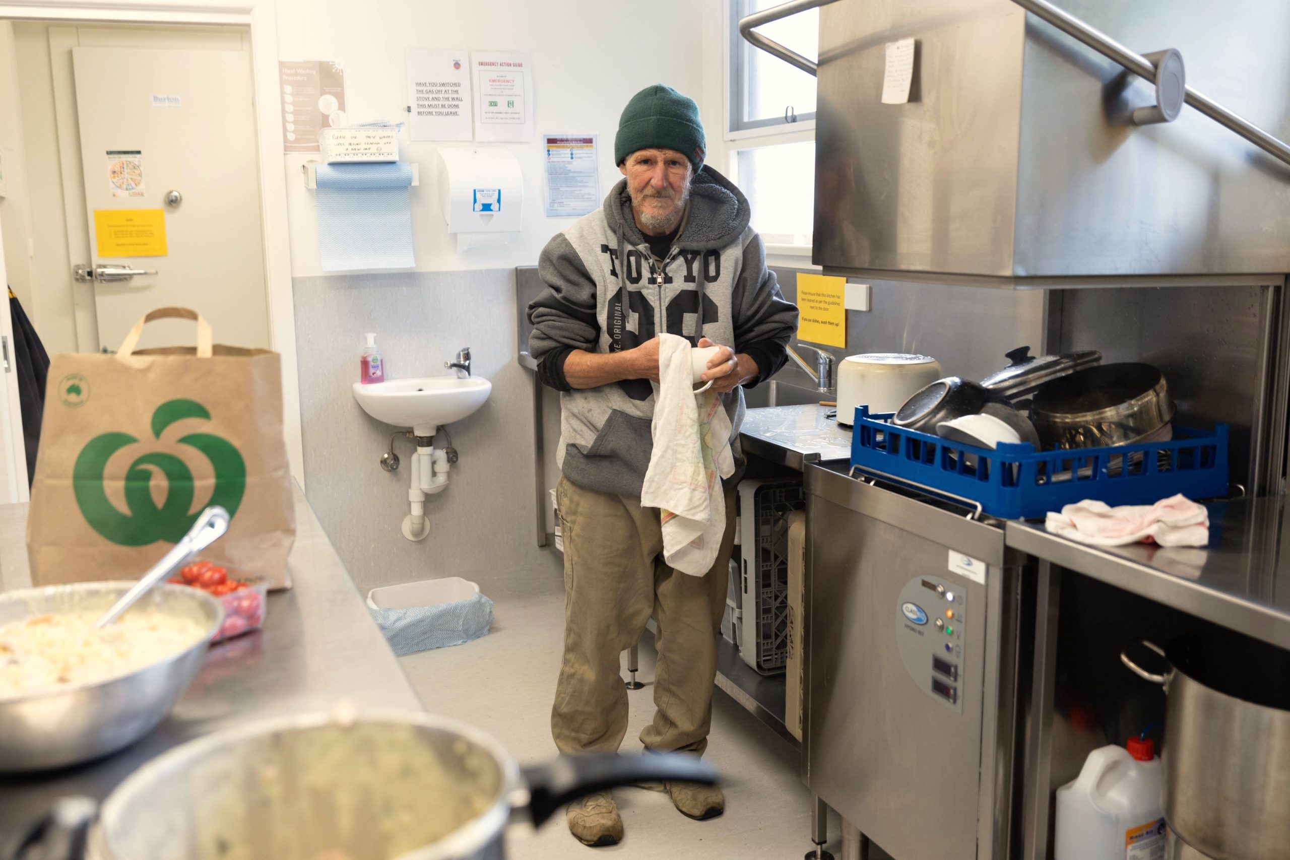 An elderly man wearing a beanie and hoodie stands in a commercial kitchen, drying dishes. A bag of groceries and a container of salad are in the foreground. The room has stainless steel appliances and shelves.