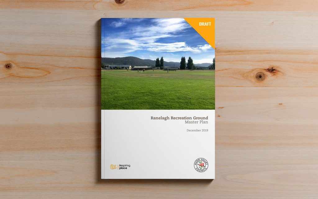 A booklet titled Ranelagh Recreation Ground Master Plan December 2019 is on a wooden surface. The cover features a landscape photo of a grassy field with trees and hills under a blue sky. There is a yellow DRAFT label at the top.