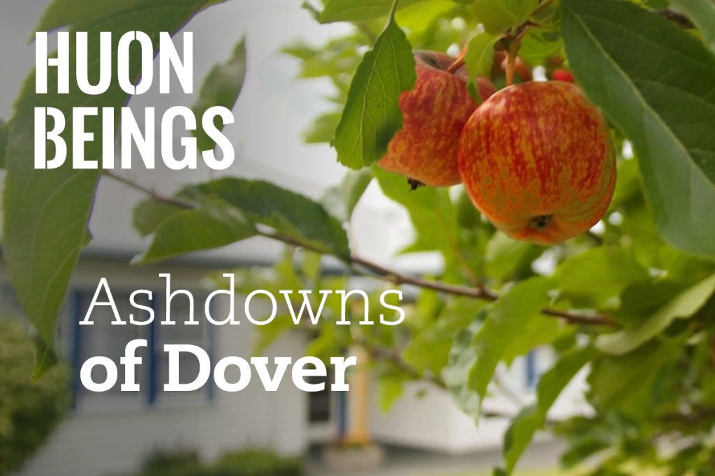 Close-up of apples hanging from a tree branch with green leaves in the foreground. A house is blurred in the background. The text reads, HUON BEINGS Ashdowns of Dover.