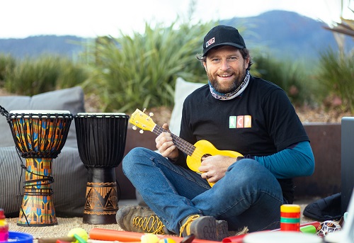 A man sits cross-legged outdoors, smiling and playing a yellow ukulele. He wears a cap and casual clothes. Beside him are colorful drums and percussion instruments. The background features lush greenery and a mountain.
