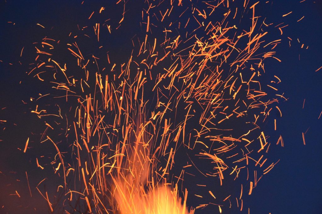 Bright orange and yellow sparks fly upwards against a dark blue night sky, creating a dynamic and energetic pattern.